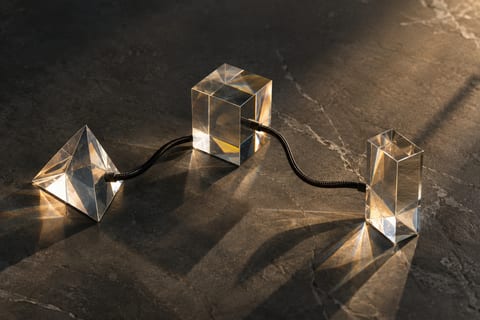 Three clear crystal prisms linked by black cables sit on dark stone, casting light flares and long shadows.