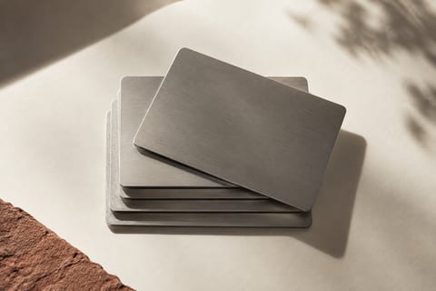 A stack of brushed metal plates lies on a pale surface under leafy shadows, beside the edge of a reddish stone.