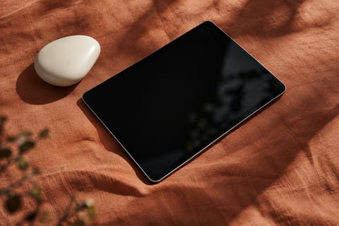 A tablet with a dark screen and a smooth white pebble lie on terracotta linen dappled with leaf shadows.